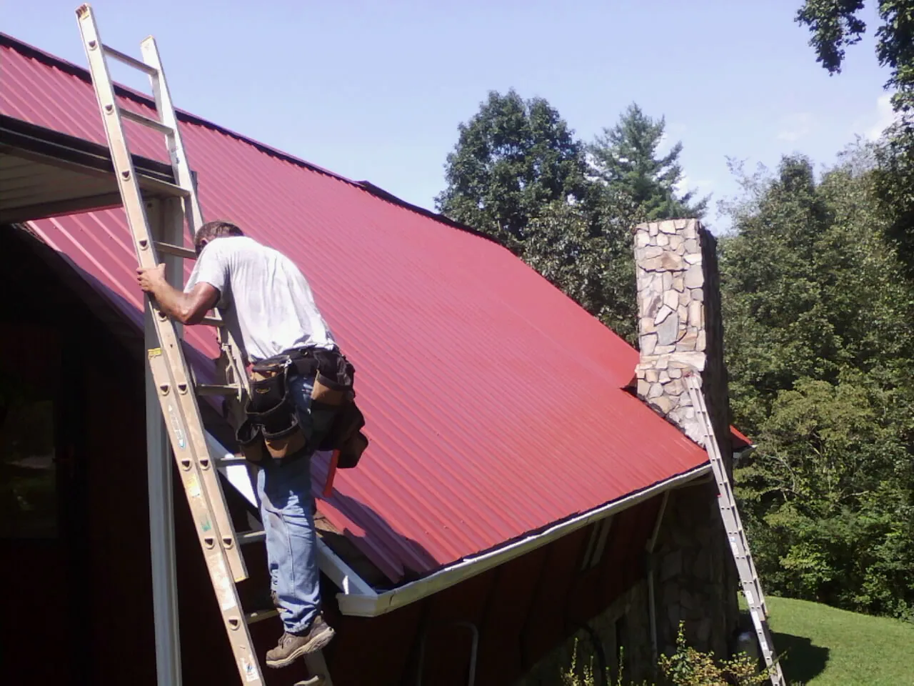 Roofer on a ladder beside a red metal roof and stone chimney — Soffit & Fascia Repair by Roofing Brothers in Fairview